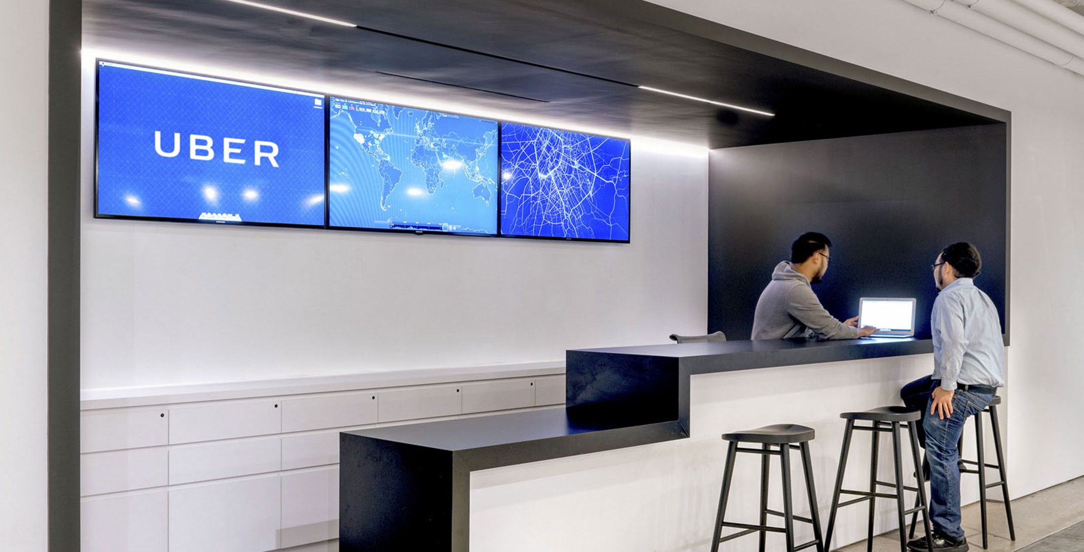 Two male employees seated at IT Support desk, with TV screens displaying global Uber map